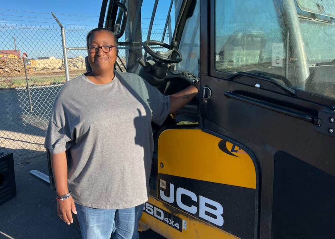 A woman stands next to the yellow cab of a forklift. they are outside.