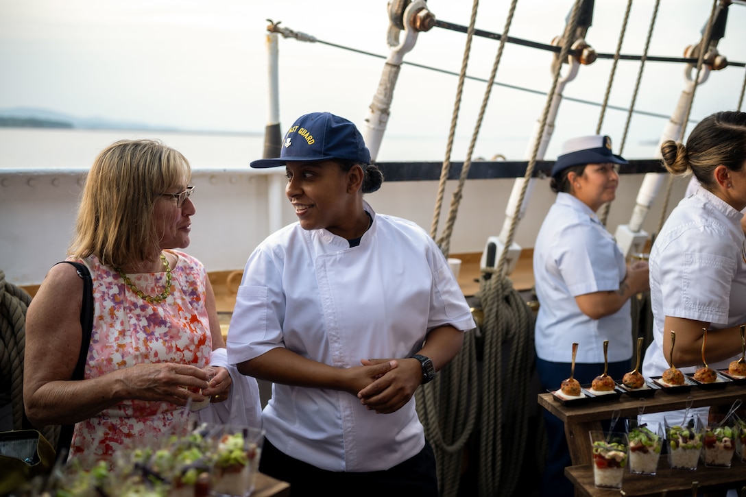 U.S. Coast Guard Petty Officer 2nd Class Fallon Ferguson, a culinary specialist assigned to Coast Guard Cutter Eagle (WIX 327) shares a laugh with a guest at Eagle’s reception to honor Rockland, Maine, as an official Coast Guard City, in Rockland, Maine, Aug. 2, 2024. Rockland recertified to become a Coast Guard City, demonstrating a mutually beneficial relationship between the local Coast Guard community and the City of Rockland.