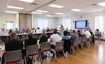 Group of people seated at tables facing each other listen to woman briefing