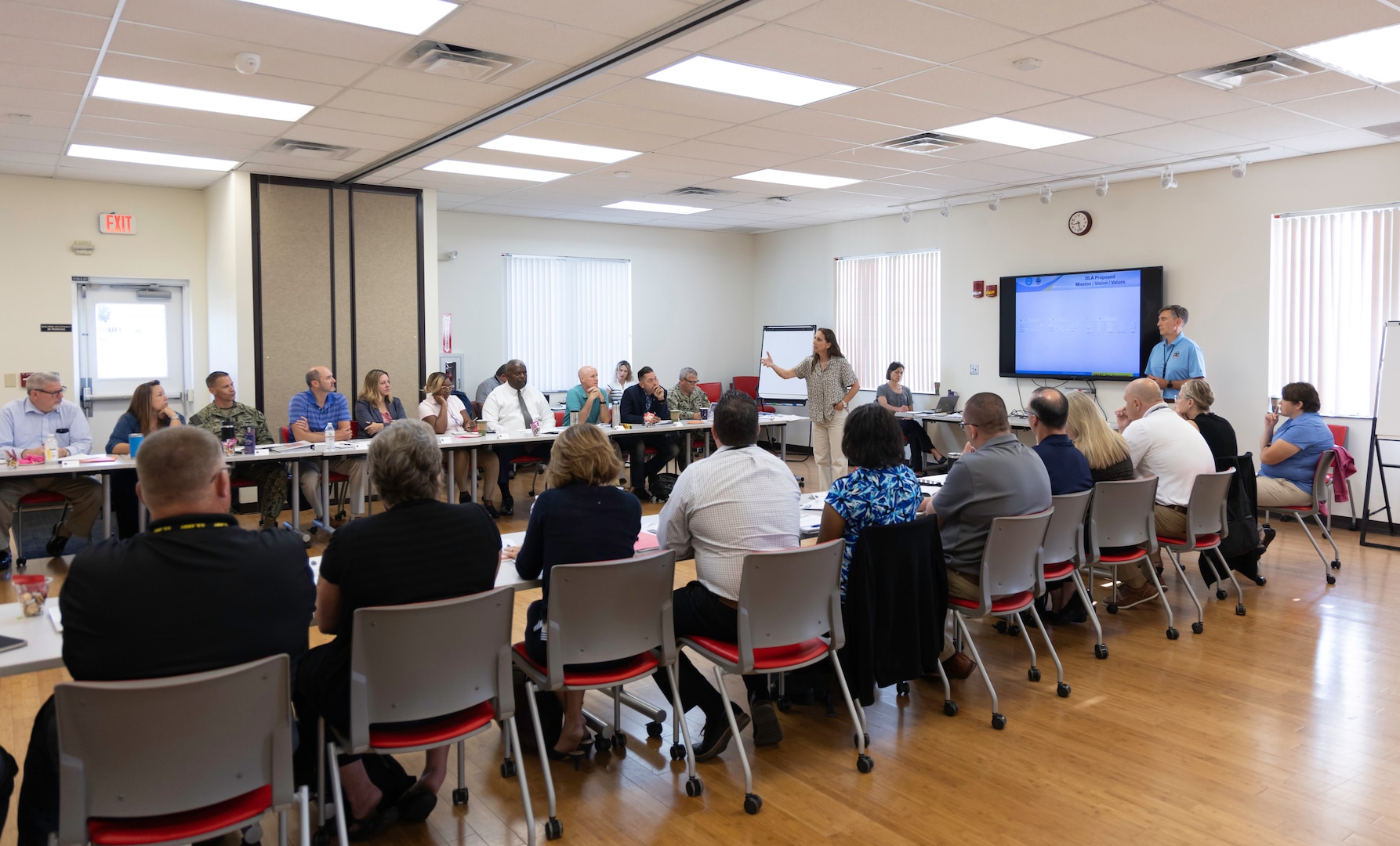 Group of people seated at tables facing each other listen to woman briefing