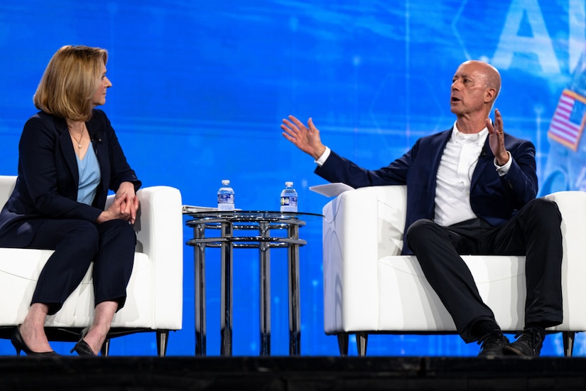 Two people in business attire speak to one another while seated on a stage.