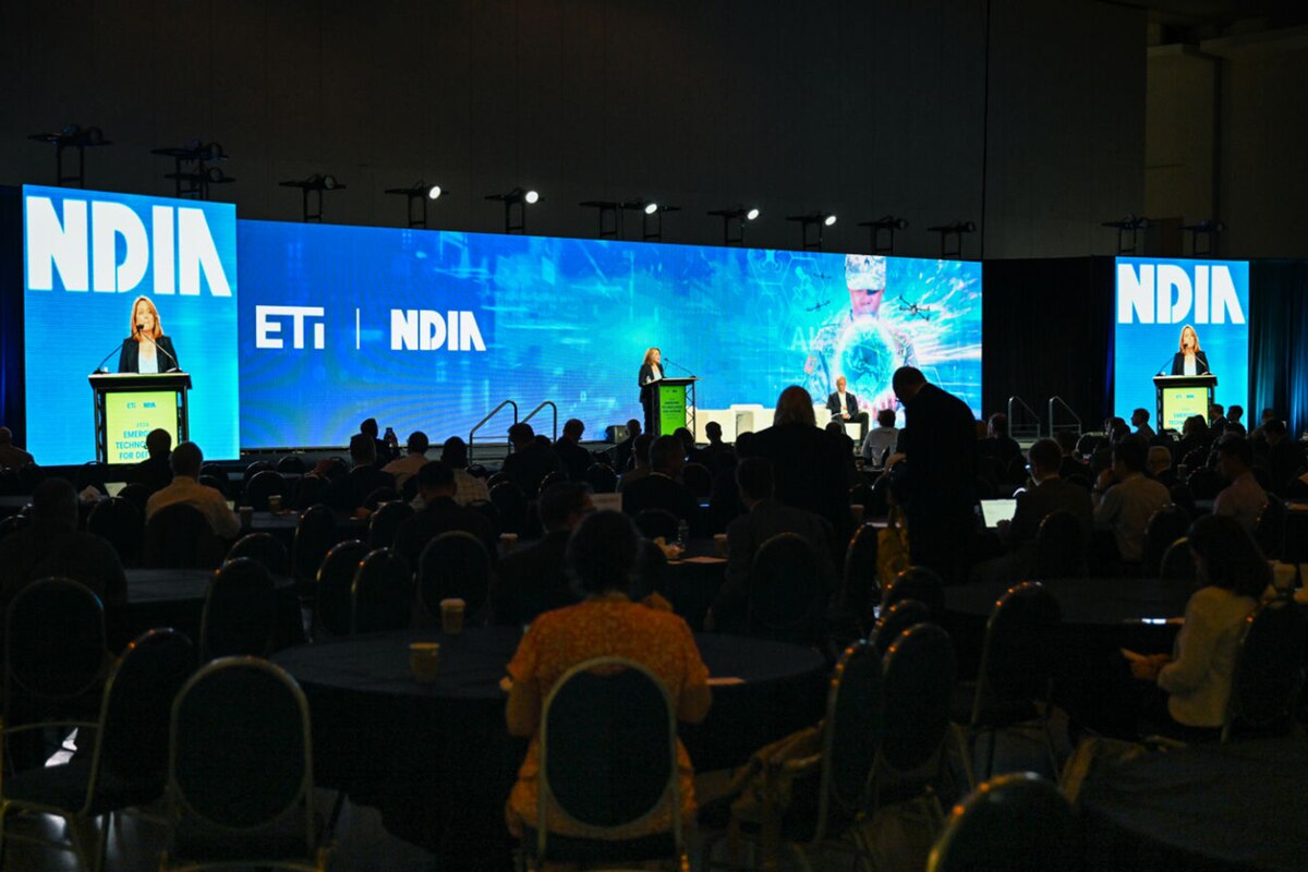 A person in business attire speaks from a podium before a large crowd in a darkened room.