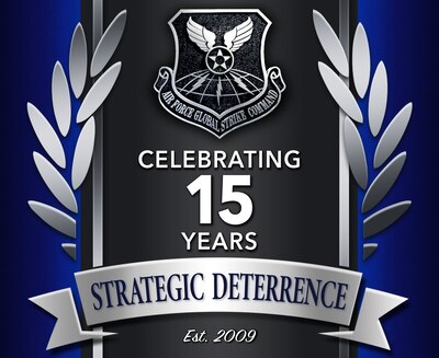 A graphic for the 15th anniversary of the founding Air Force Global Strike Command. The graphic features a dark blue textured background with a dark gray stripe in the middle of the screen that runs from top to bottom. There are silver wings on either side of the dark gray stripe. The very center of the design shows a silver and metallic looking version of the AFGSC shield. Below the shield are the words, "Celebrating 15 years" in white font. Underneath those words, on a horizontal silver banner is the text, "Strategic Deterrence" in a dark blue font. Underneath the banner is the text, "Est. 2009" in a white script font.