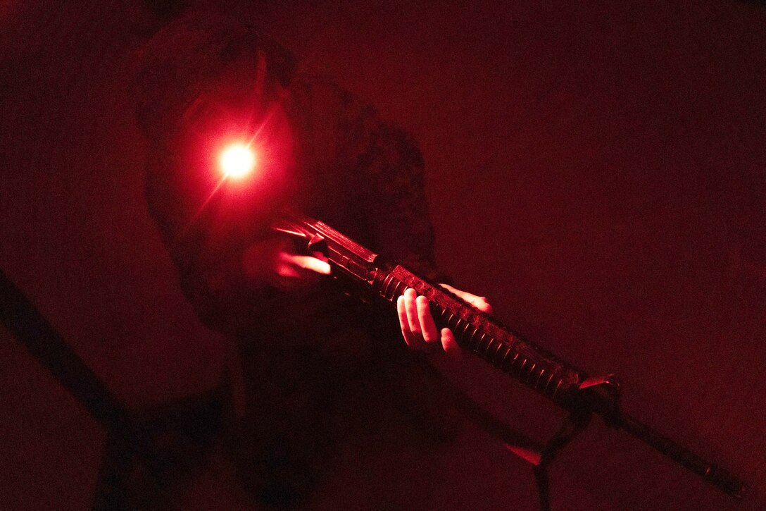 A Marine carrying a weapon uses a helmet-mounted light to see in a dark room under red lights.