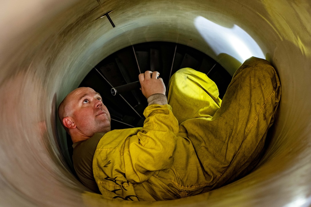 An airman in yellow overalls lies curled in the interior of an aircraft holding a flashlight to perform an inspection.