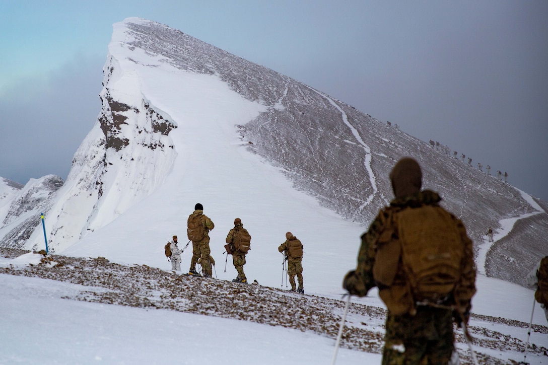 Marines climb a snow-covered mountain on a cloudy day while others stand below and watch.