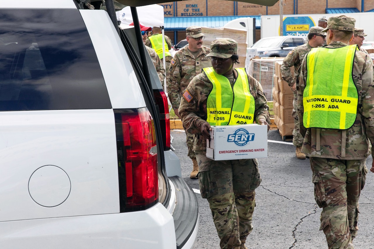 A soldier carries a box labeled Emergency Drinking Water next to an open SUV with more soldiers milling about outside in the background.