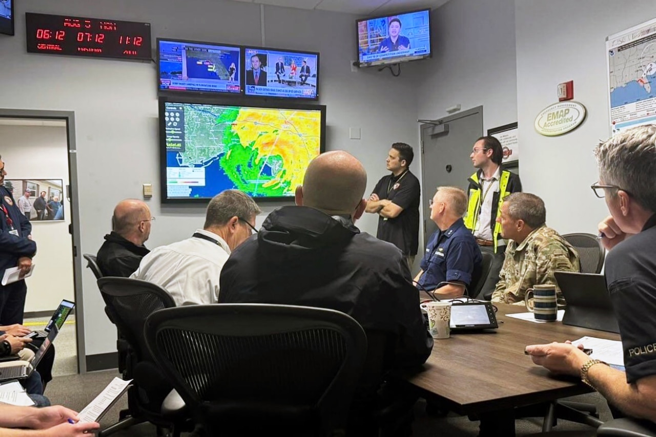 A group of workers sit around a table looking at a TV displaying a hurricane forecast as more TVs line the walls.