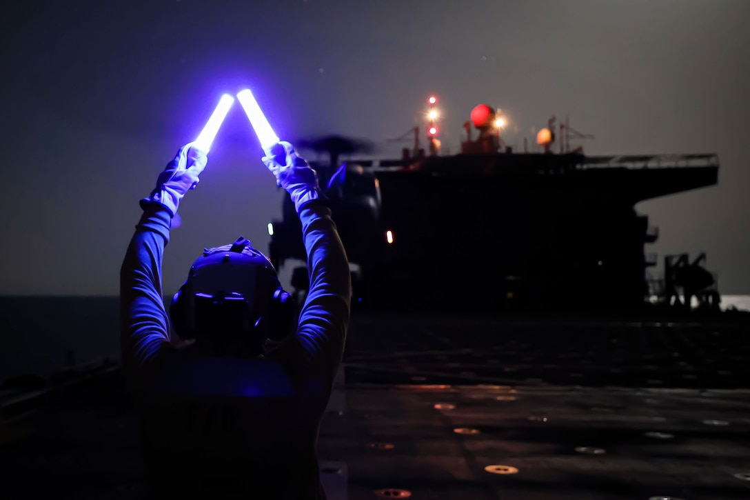 A sailor uses aircraft marshalling wands to signal a military helicopter on the flight deck of a ship at night.