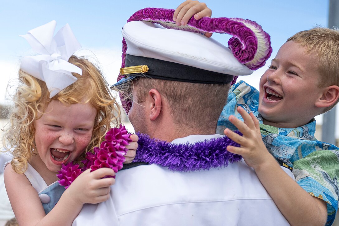 A sailor wearing floral leis holds two smiling children.