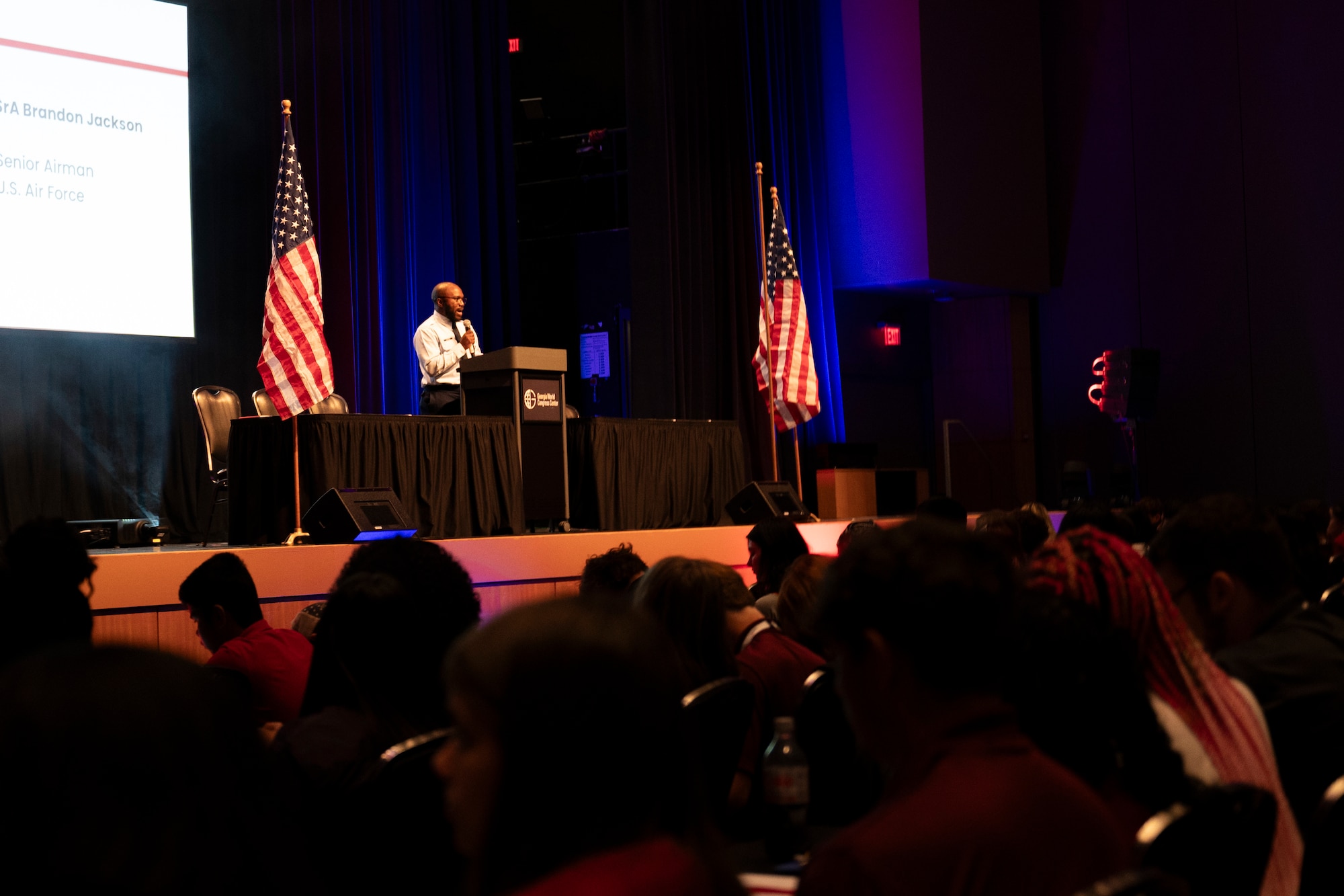 A man stands on stage speaking to a crowd in a dimly lit room.