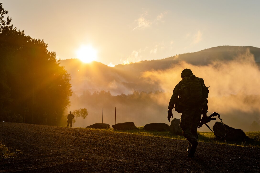 A soldier carrying a weapon walks up a hill as sun rays beam from behind a mountain as a fellow soldier watches.