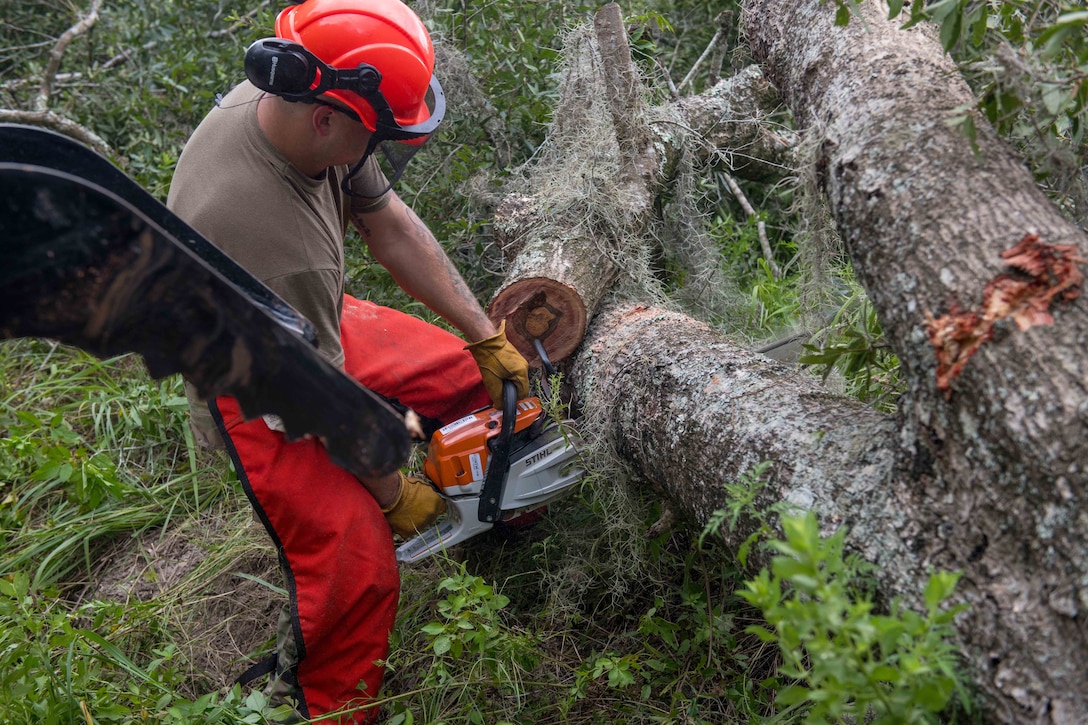 An airman wearing protective gear uses a chainsaw to cut fallen trees.