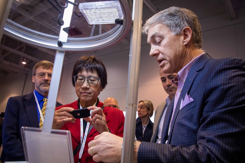 A woman stands next to a man holding a small device. Both are wearing business attire.