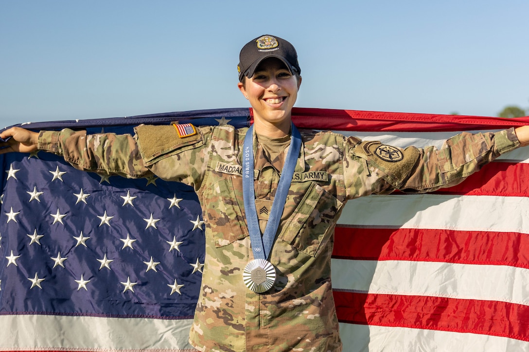 A soldier with a silver medal slung around their neck poses while holding the U.S. flag behind them.
