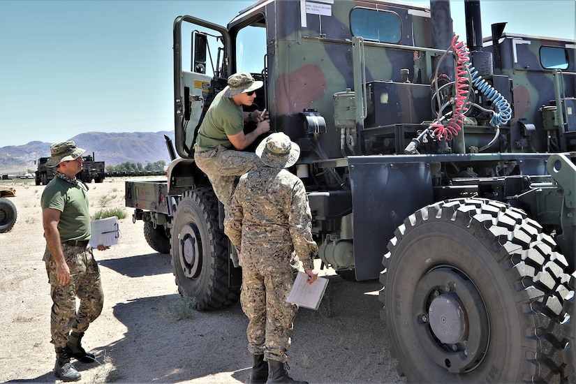 Military personnel stand near the driver-side door of a large military vehicle. One individual has stepped up on the vehicle.