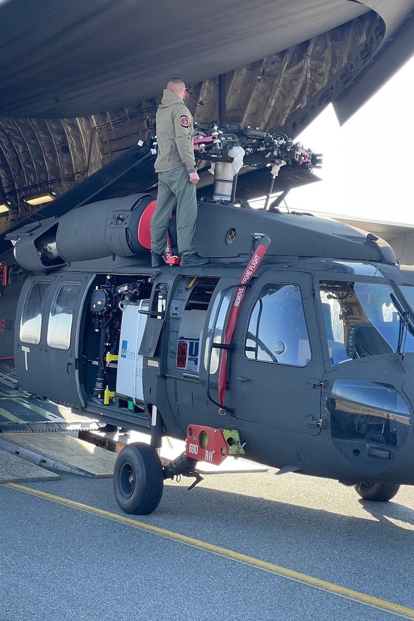 A person stands on top of a helicopter which is rolling down a ramp extending from inside an aircraft.