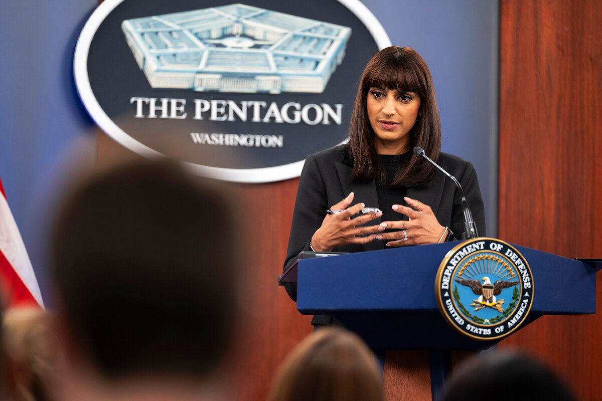 A woman stands at a lectern with her hands gesturing inward. A sign that says, The Pentagon is behind her.