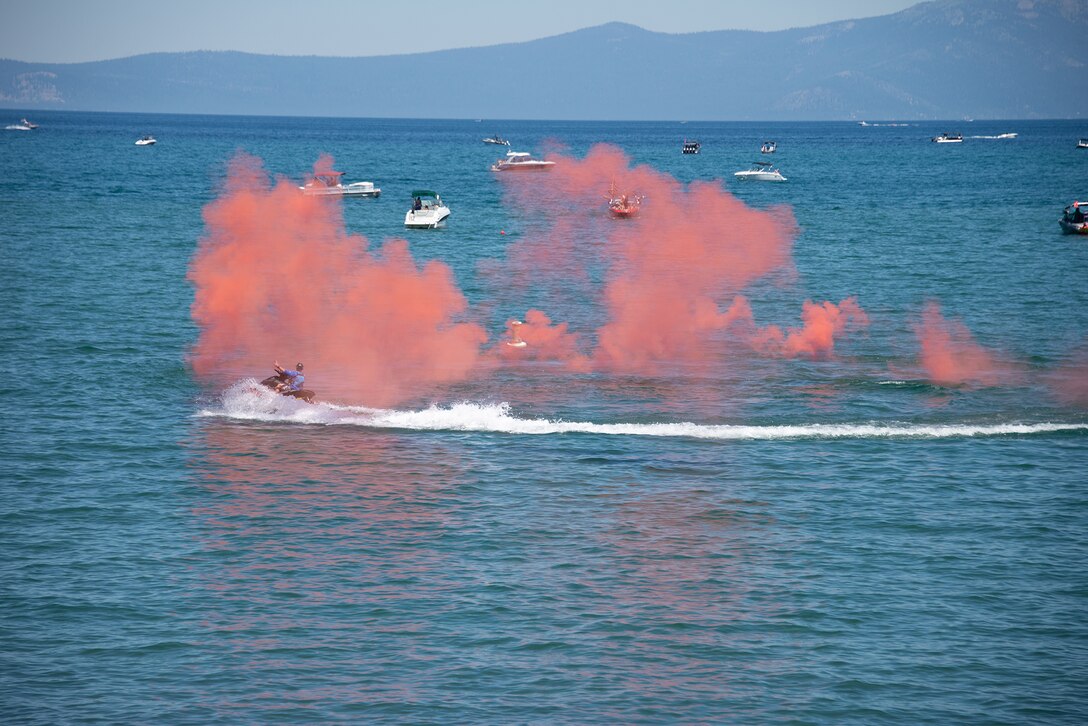 A person rides a jet ski in the foreground as pink smoke hovers over the water during daylight. Small boats are visible in the background.