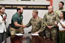 A man in a dark green polo shirt (on left) is shaking hands with a man in Army camouflage (on right) while beginning to stand up from chairs at a glossy, dark brown conference table.