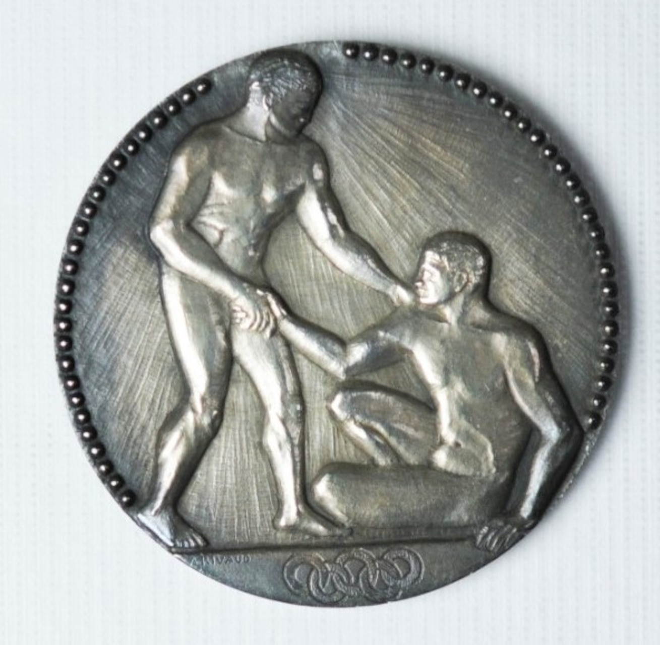 On the obverse stands a nude victorious athlete, taking the hand of his defeated rival, assisting him to rise from his position on the ground. Under the image appears the Olympic rings