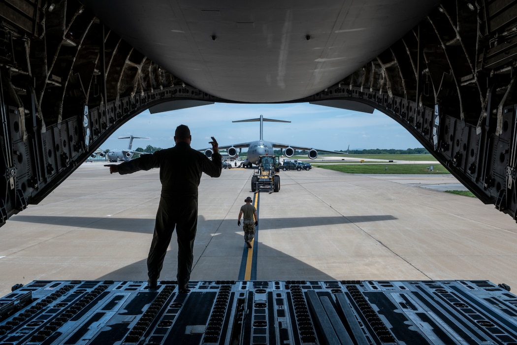 A man stands in the back of an open cargo area of an aircraft as he directs equipment.