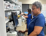 Two pharmacy technicians stand in front of computer screens.