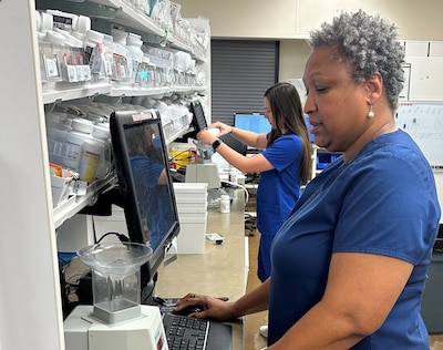 Two pharmacy technicians stand in front of computer screens.