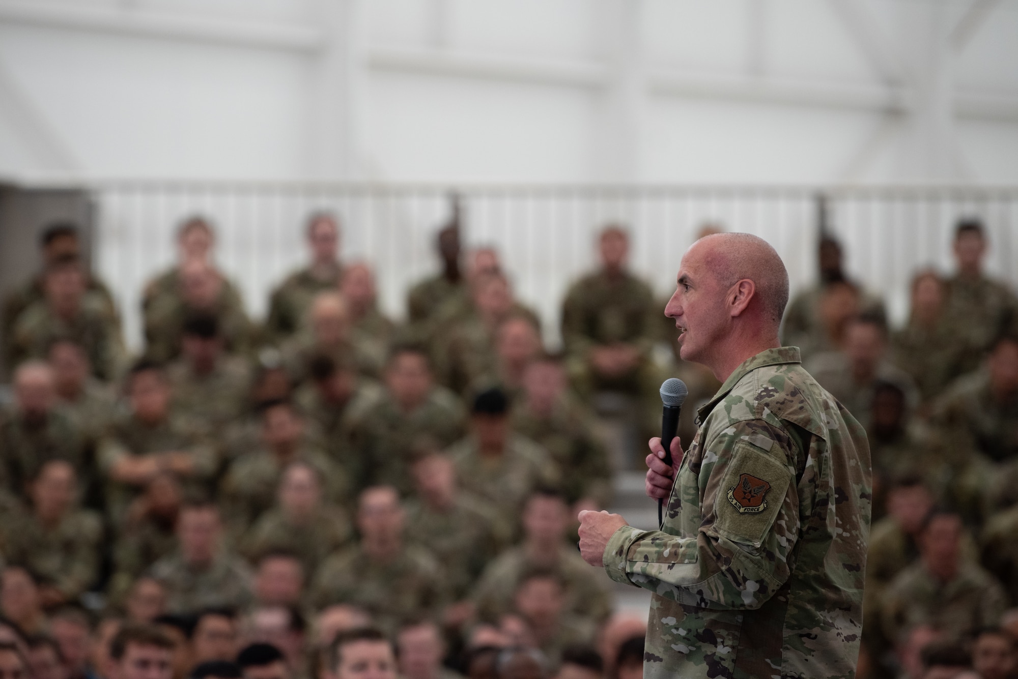 A man in military uniform uses a microphone to address a crowd of people seated in a military hangar.