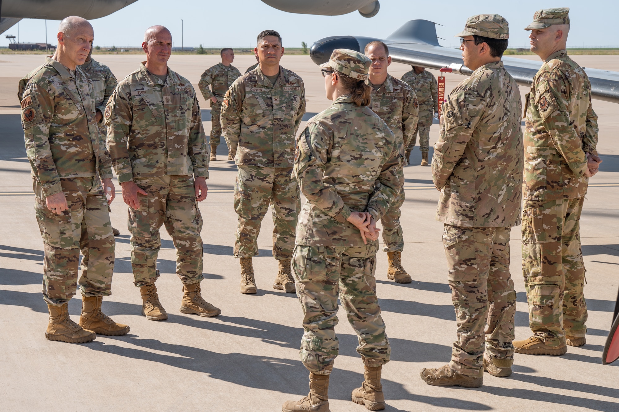 Three individuals in military uniform stand on a military tarmac and address a small group of men in military uniform who are standing in front of a military aircraft.