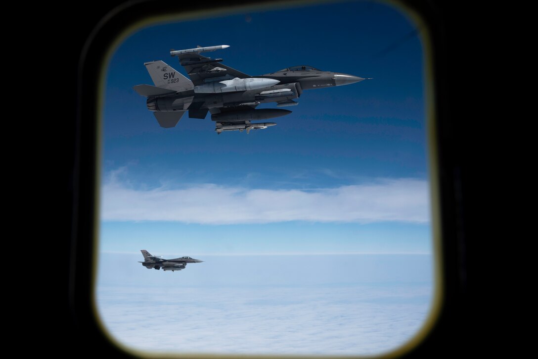 Two aircraft fly next to each other near clouds as seen through the window of another aircraft flying nearby.