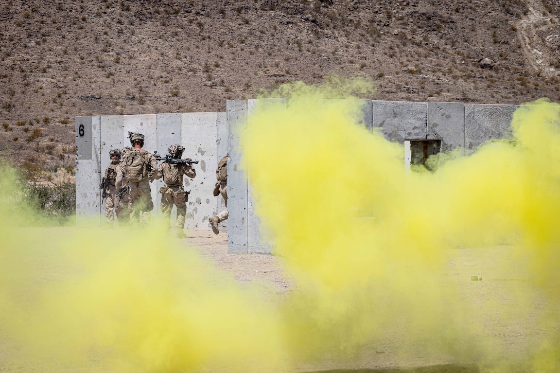 Marines carrying weapons move around concrete structures in front of a mountain as clouds of yellow smoke rise in the foreground.