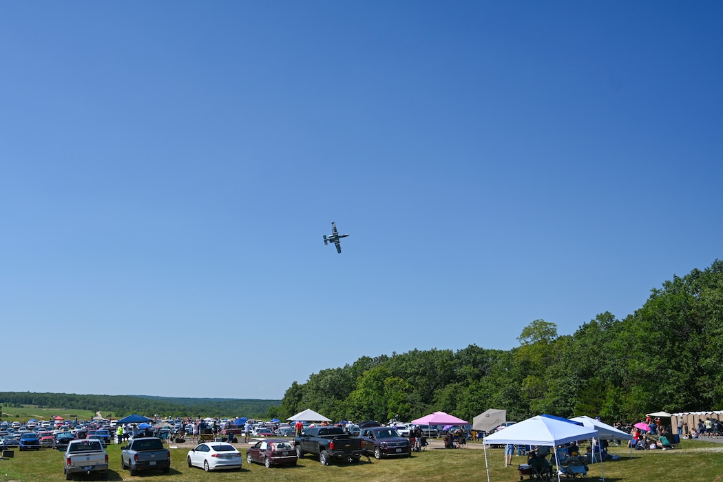 An A-10 banks over a crowd of people under sun shade canopies.