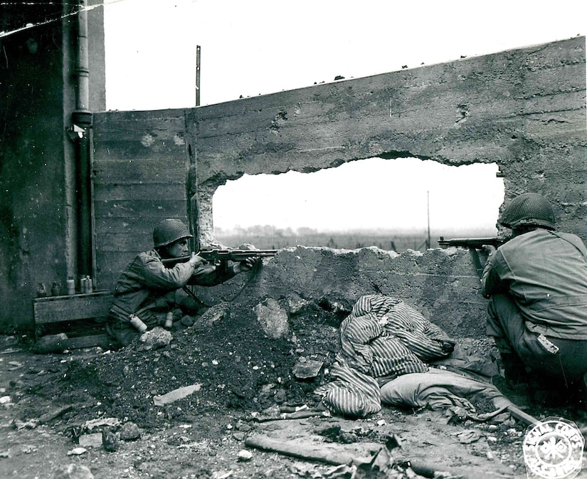 Two crouching men aim their guns through a blasted-out hole in the wall of a cement barrier.