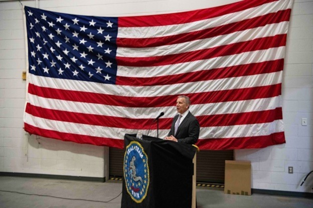 A man wearing business attire stands behind a podium in front of a large American flag.