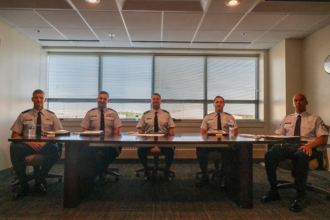 Five men in dress uniforms sit behind a long table.