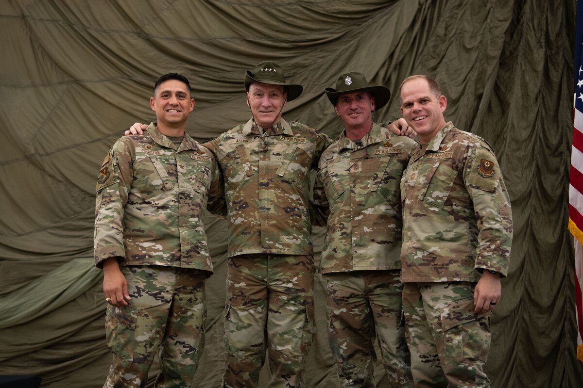Four men in military uniform pose for a photo on a stage. The two men in the center are wearing hats.