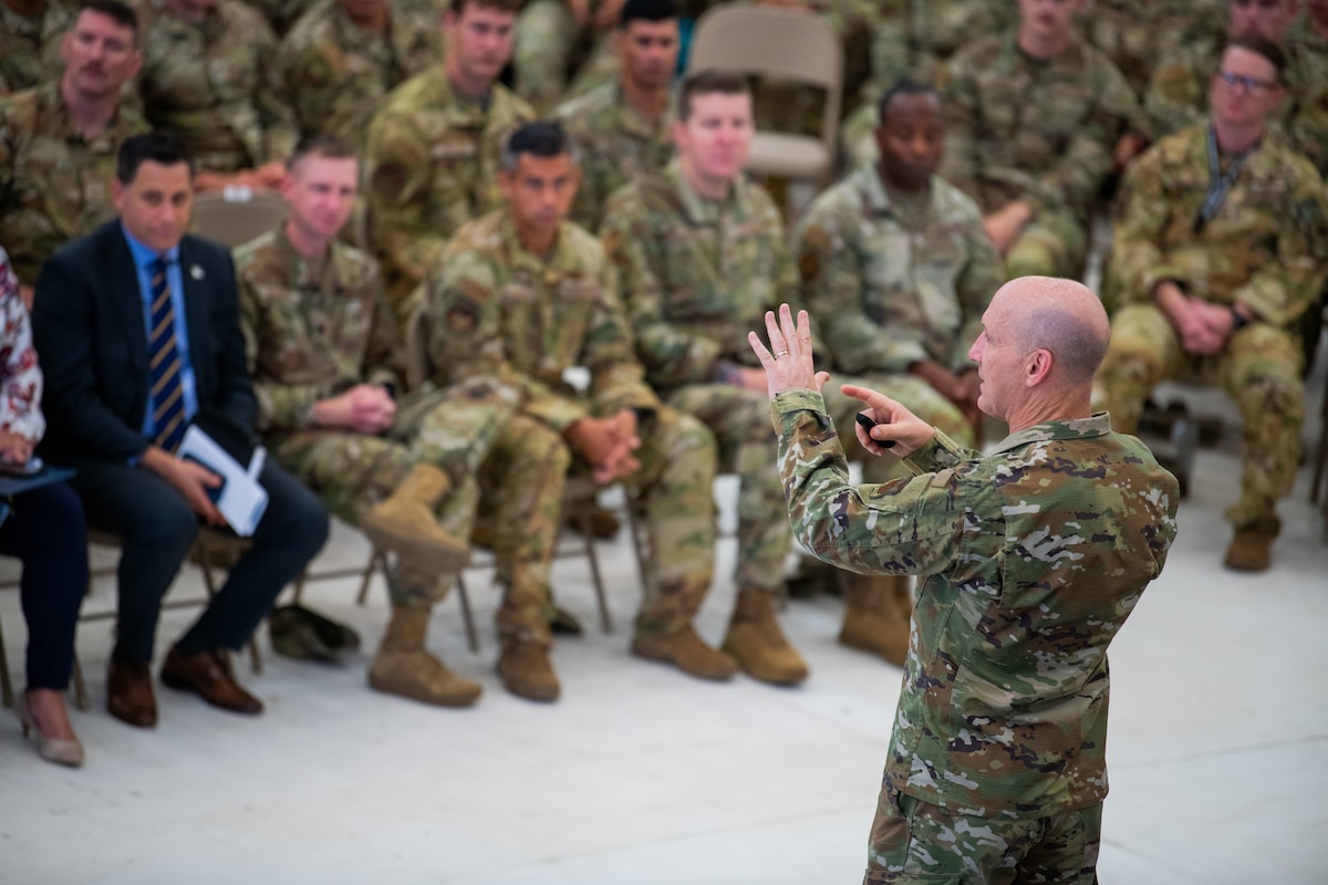 A man in military uniform stands on a stage inside a building addressing a seated crowd of people in military uniform and business attire.