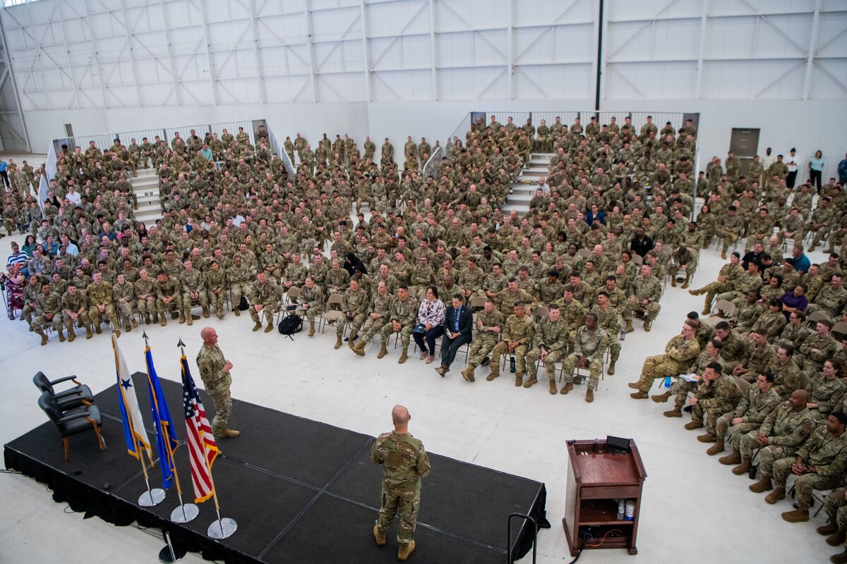 Two men in military uniform stand on a stage inside a hangar addressing a seated crowd of people in military uniform.
