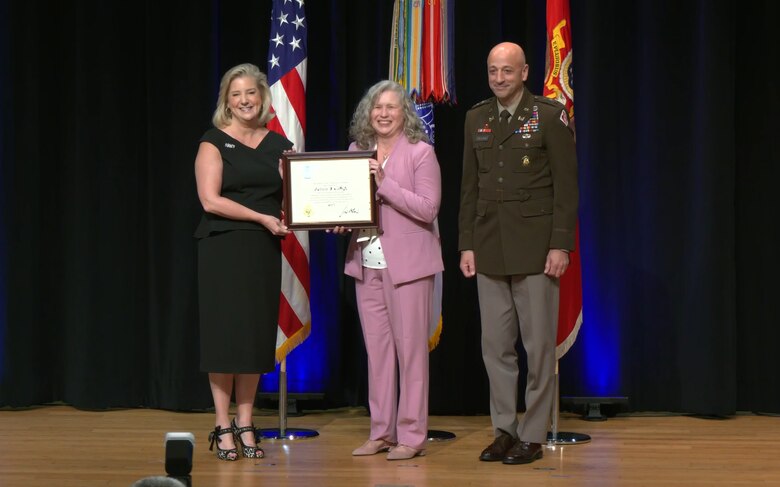 Three people on a stage. One woman on the left side is handing another woman in the center an award.