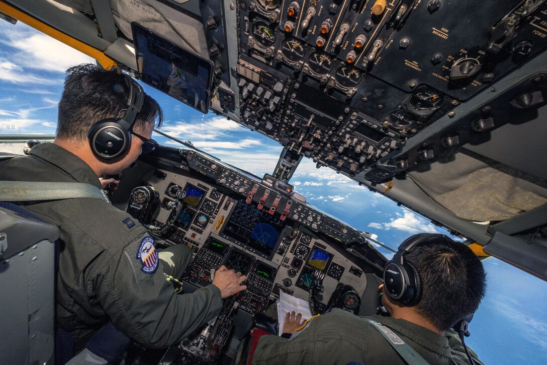 Two pilots in an aircraft cockpit focus on instruments and controls, with a blue sky visible through the front windows.