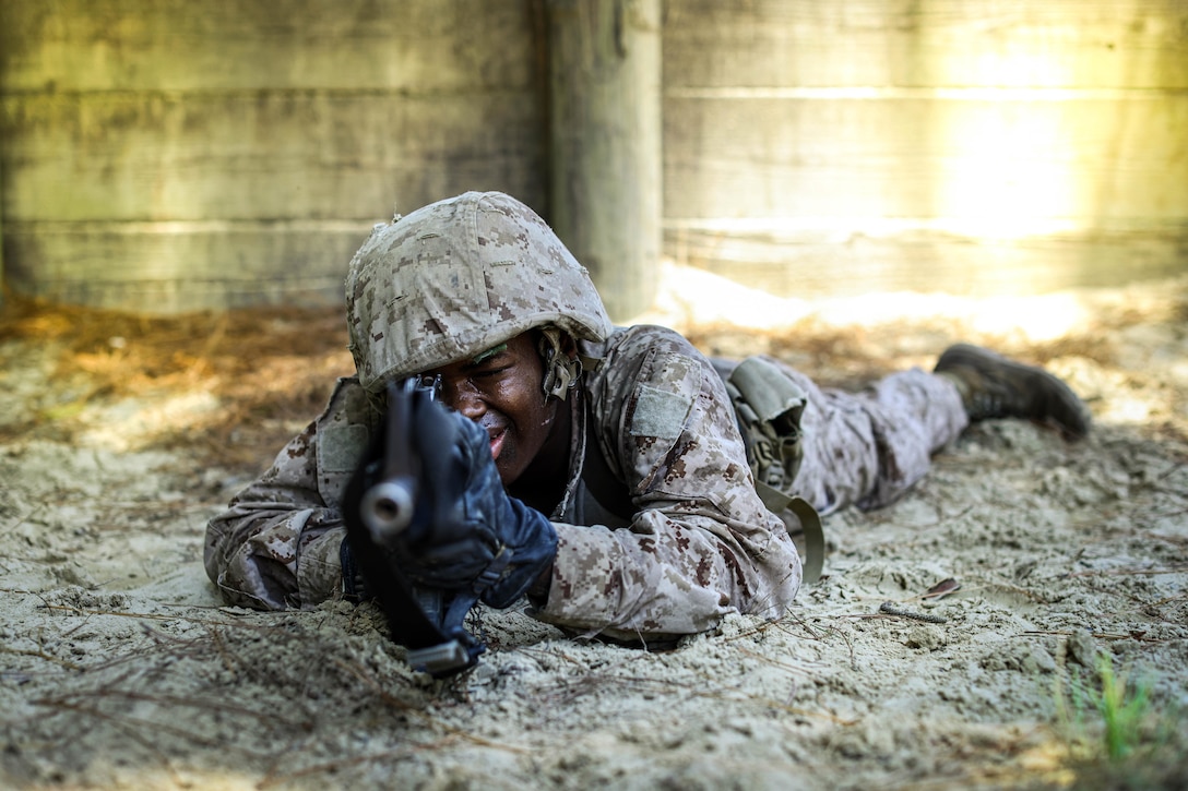 A service member recruit in camouflage lies on the ground aiming a firearm surrounded by a sandy environment and wooden structures.