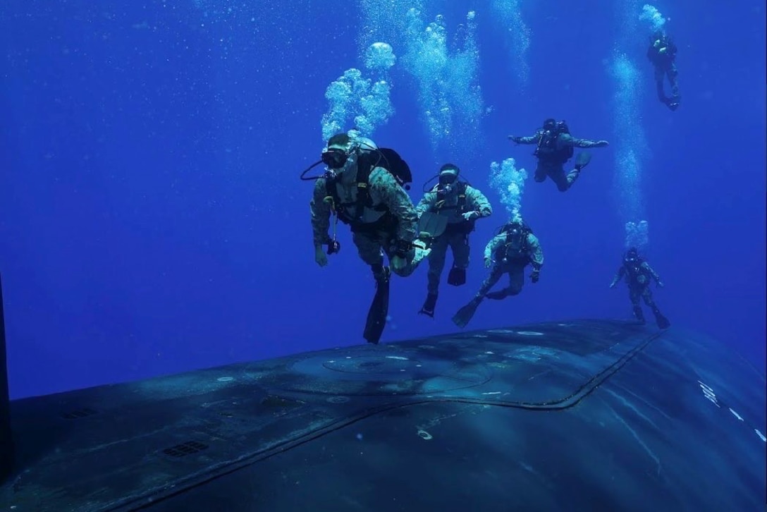 Military divers in full gear swim above a submerged submarine in deep blue waters.