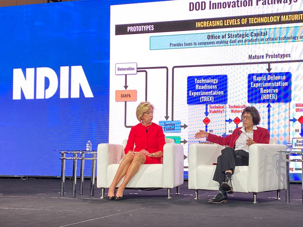 Two women sit in front of a large screen with a technical presentation on it.