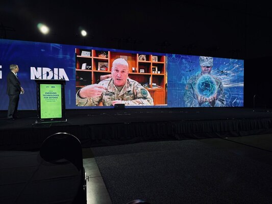 A large screen in a dark room is broadcasting a man in a camouflage uniform sitting at a desk with a wooden bookshelf behind him. To the right of the large screen is an illustration of a woman in a camouflage uniform holding a ball of energy. To the left, a man stands next to the lectern looking at the large screen.