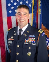 Army Soldier smiling while in ceremonial uniform in front of the U.S. flag and the Army flag.