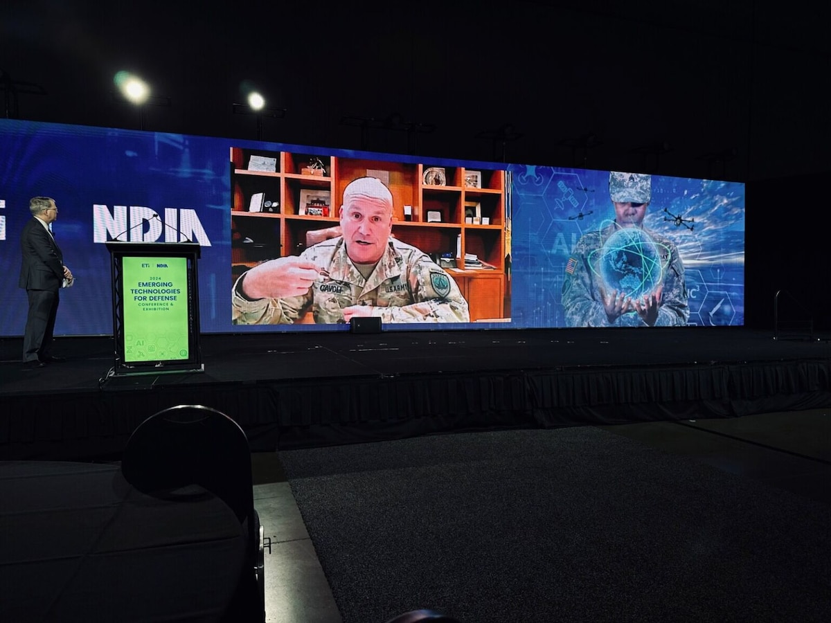A large screen in a dark room is broadcasting a man in a camouflage uniform sitting at a desk with a wooden bookshelf behind him. To the right of the large screen is an illustration of a woman in a camouflage uniform holding a ball of energy. To the left, a man stands next to the lectern looking at the large screen.