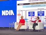 Two women sit in front of a large screen with a technical presentation on it.
