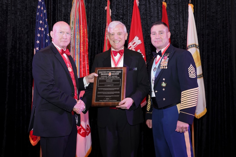 Three men stand in front of flags, one man on the right is  wearing an Army dress uniform, the other two men are wearing suites.