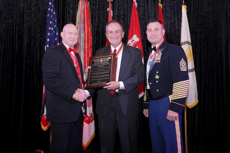Three men stand in front of flags, one man on the right is  wearing an Army dress uniform, the other two men are wearing suites.